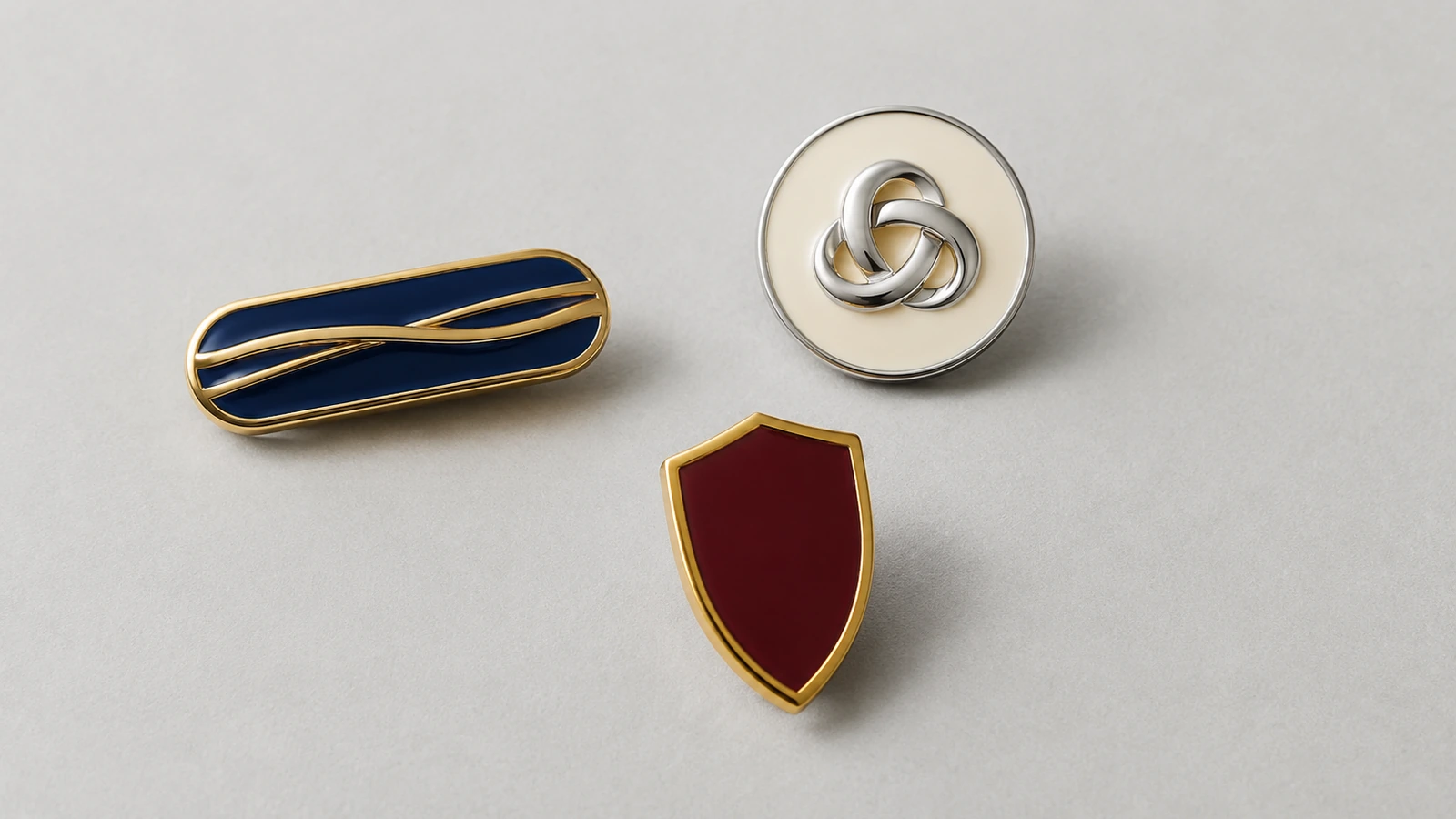 Minimalistische unbedruckte Pins mit glatten, erhabenen Metallbändern und einer ineinandergreifenden Schlaufenform, die Typografie andeuten, ohne lesbare Zeichen