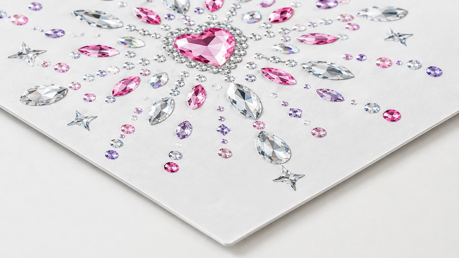 Nahaufnahme eines rosa Strassmotivs mit Herzmitte und klaren, rosa und lila Steinen auf einem weißen Bogen